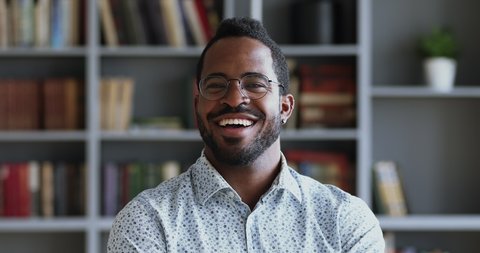 Happy african young adult businessman wear optical glasses dental smile look at camera in home office, mixed race student professional teacher posing on bookshelves background, close up portrait