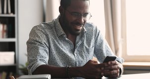 Smiling young adult african american guy student worker using mobile apps sit at home office table, happy mixed race male user enjoying browsing online social media study online on modern tech gadget