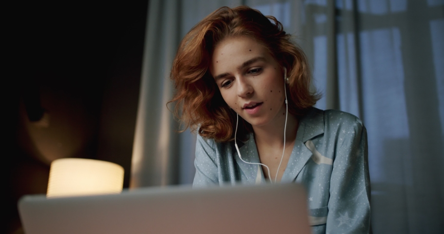Low portrait shot of cheerful female smiling staying online in social media in the evening, happy teen girl laughing watching YouTube video using laptop and earphones at home dressed in trendy pajama Royalty-Free Stock Footage #1043984488