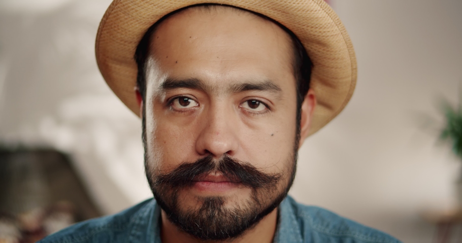 Close up of super surprised emotional Hispanic male with mustache and beard indoor in hat, cheerful brunette guy smiling of unexpected news showing expressive facial expression dressed in jeans shirt | Shutterstock HD Video #1043987641