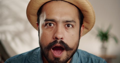 Close up of super surprised emotional Hispanic male with mustache and beard indoor in hat, cheerful brunette guy smiling of unexpected news showing expressive facial expression dressed in jeans shirt