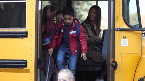 Smiling mixed race female driver of school bus waving goodbye to elementary age kids going to school. Diverse schoolboys and schoolgirls taking turns leaving yellow bus arriving to school