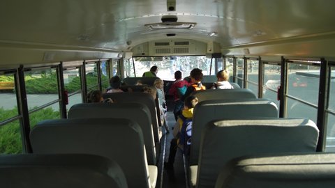 Back view of elementary age schoolchildren rising from seats to get off school bus. Mixed race woman driver waving goodbye to diverse preadolescent kids leaving school bus and going to school
