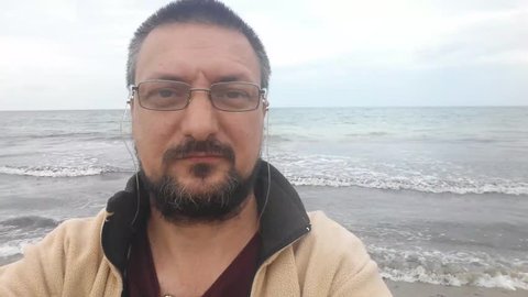 Bearded adult man with glasses taking selfie selfportrait in video mode on Black Sea shore.
