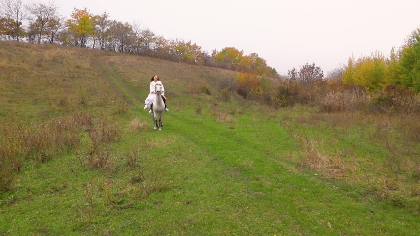 Long-haired girl in white dress is riding horse along forest
