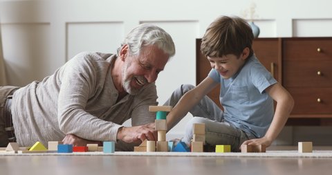 Happy old senior grandfather teach little grandson playing with wooden blocks on floor, carefree two generation family grandparent and cute small grandchild having fun building tower laughing at home