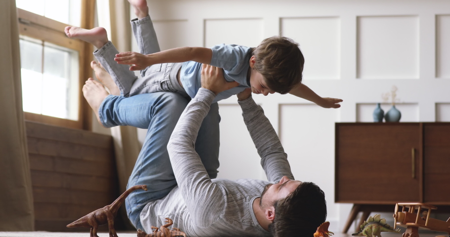 Carefree loving family adult dad hold cute little kid son pretend superhero plane having fun lying on floor, happy father lifting small child boy up flying enjoy lifestyle playing funny game together Royalty-Free Stock Footage #1044003337
