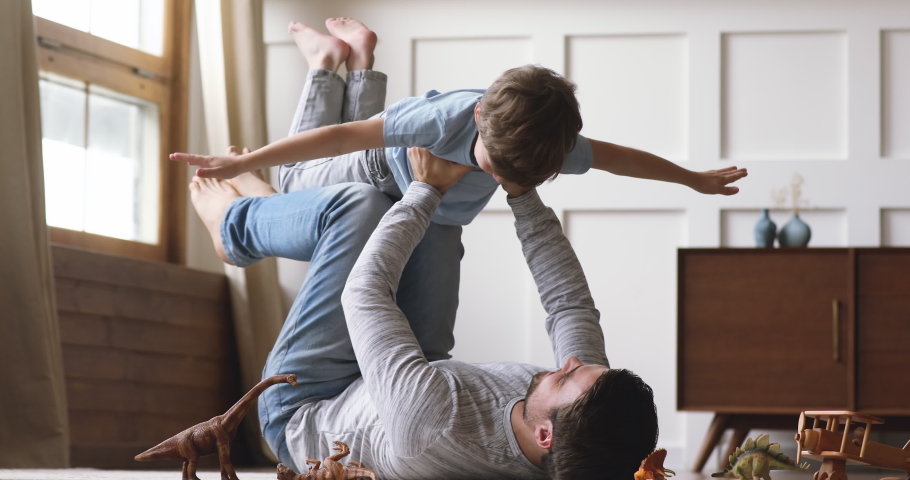 Carefree loving family adult dad hold cute little kid son pretend superhero plane having fun lying on floor, happy father lifting small child boy up flying enjoy lifestyle playing funny game together | Shutterstock HD Video #1044003337