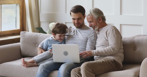 Friendly three 3 age multi generation men family using laptop together, happy old grandpa, young adult grown son father and kid boy grandson laugh watch online tv show on computer at home sit on sofa