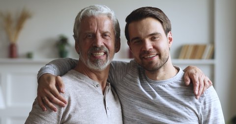 Happy handsome two age generations men family posing for portrait at home, smiling young adult son hugging senior older grey haired father embracing together looking at camera, close up portrait