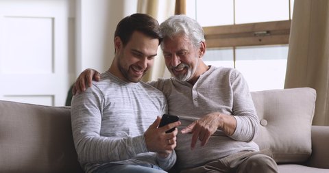 Happy two generations family old man father embrace young adult son holding smart phone discuss social media online news new mobile app having fun talking enjoying using cellphone at home sit on sofa