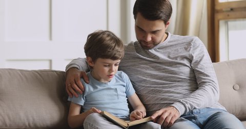 Caring single parent adult dad teach focused concentrated preschool child son reading book learning education fairy tale story bonding spend free time enjoy family lifestyle hobby at home sit on sofa
