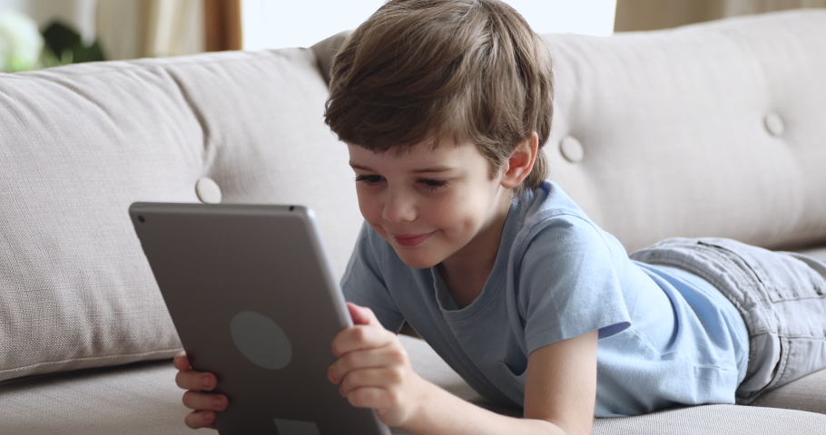 Happy cute preschool boy using tablet lying on sofa, smiling small kid watch online cartoons on pad computer in internet having fun play game on tab alone at home, children and digital tech concept Royalty-Free Stock Footage #1044003400