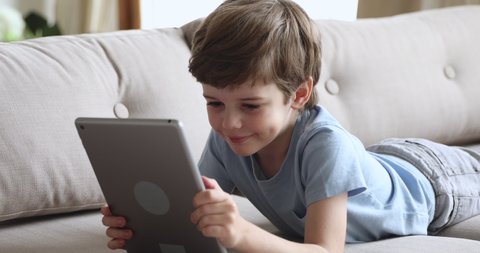 Happy cute preschool boy using tablet lying on sofa, smiling small kid watch online cartoons on pad computer in internet having fun play game on tab alone at home, children and digital tech concept
