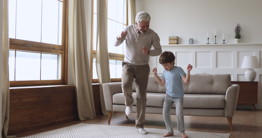 Happy two age generations active family dancing in living room, carefree old senior adult grandfather and cute preschool grandson having fun listening music jumping enjoying time together at home Royalty-Free Stock Footage #1044003406