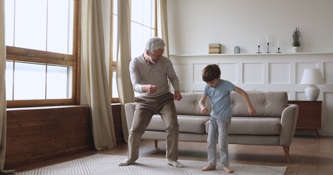 Happy two age generations active family dancing in living room, carefree old senior adult grandfather and cute preschool grandson having fun listening music jumping enjoying time together at home