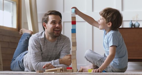 Happy adult parent dad helping cute preschool child son playing toys together on floor, caring father and little kid boy having fun laugh building constructor tower of colorful wooden blocks at home