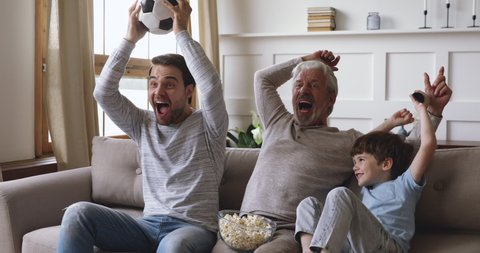 Excited three generation men family grandson young father and old grandfather fans hold soccer ball remote control watch tv sport game celebrate goal television team victory in tv match sit on sofa