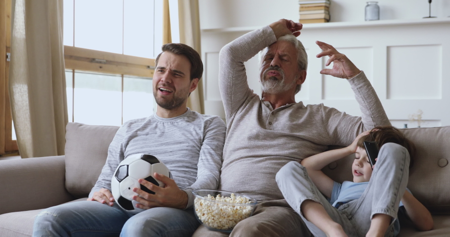 Frustrated disappointed three generations male family old grandpa, young son father and kid grandson fans supporters hold soccer ball watch tv sad by team losing football match sport game sit on sofa Royalty-Free Stock Footage #1044003445