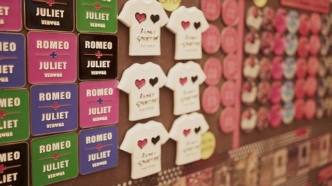 VERONA, ITALY - JUNE 2019: Handheld pan shot of rows of souvenir magnets of Romeo and Juliet in Juliet s House, Verona. Handheld shot of magnets of different colours in shape of hearts