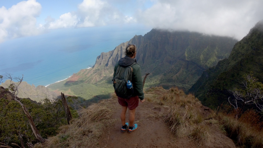 Man enjoys views of mountain and ocean from hike lookout point. Young man hiking in Hawaii. Na Pali trail