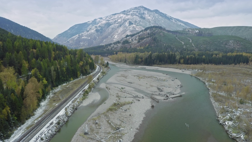 Aerial: highway surrounded by forest, mountains covered in snow. Alongside is the Middle Fork Flathead River. Glacier National Park, Montana, USA 