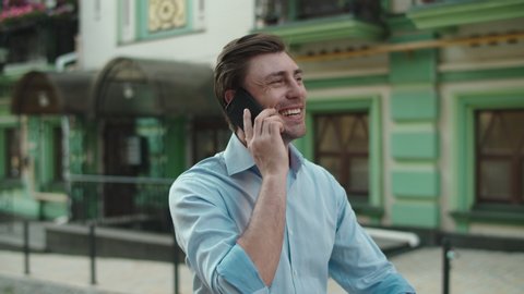Young business man walking with mobile phone at street. Smiling businessman call phone outdoors. Cheerful man talking phone outside. Office worker going to work