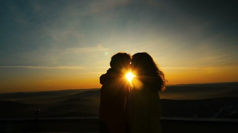 couple in love kisses standing on snowy slope at back pictorial orange sunset between heads against hilly landscape in mist