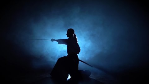 Silhouette Aikido Master Wearing Traditional Samurai Hakama Clothes Takes His Japanese Sword out of Scabbard and Swings with It, from a sitting position. He's in the Spotlight Darkness Surrounds Him.