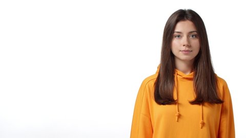 Hey might like it. Slow-motion adorable young brunette woman in orange hoodie invite to event, check out promo, pointing fingers left wink camera and show thumbs-up with smile, white background