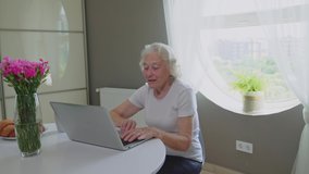 Front view of senior smiling woman having video call on laptop. Old female sitting at table with flowers and croissants in kitchen and typing on notebook. Round window behind. Concept of technology.