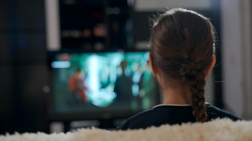 A young girl sitting on a sofa watching a movie on TV.