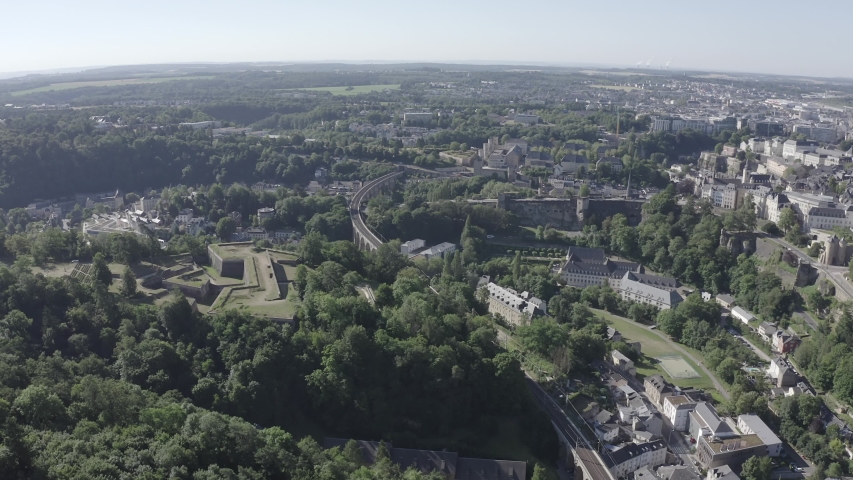 Luxembourg, Historical. The fortified old quarters of Luxembourg City rise from a steep rocky outcrop above the deep green Alzette and Petrusse valleys, with the Adolphe Bridge spanning the gorge below. The aerial camera glides over the UNESCO-listed ramparts, revealing the Bock Casemates, church spires, and dense clusters of historic buildings that earned the city its nickname as the Gibraltar of the North. Morning sunlight casts long shadows across the stone fortifications and lush tree canopy, illuminating the contrast between the medieval upper town and the verdant canyon walls. The double-arch stone bridge, once boasting the largest stone arch in the world at the time of its 1903 completion, threads through the valley floor while the old town
