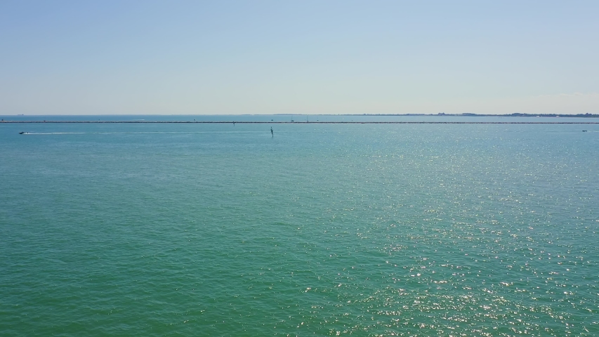 Venice, Italy. The Venetian lagoon stretches across the frame, its surface a gradient of turquoise and deep blue under a clear summer sky. The camera slowly tilts downward from an elevated position, revealing a stone breakwater of large pale rocks along the shoreline, a paved walkway, and a sandy path in the foreground. The water sparkles with reflected sunlight, and a distant low horizon line separates the calm sea from the pale sky. The lagoon is protected by barrier islands and coastal defenses that have safeguarded Venice for centuries, with sea-defenses constructed along the coastal strip since the 18th century. The scene conveys a serene, sunlit coastal atmosphere typical of the Adriatic shoreline in summer.