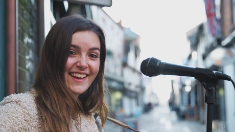 Portrait of young woman busking playing acoustic guitar outdoors in street - shot in slow motion