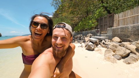 Young Multi Race Couple Making Selfie At Paradise Beach. Girl Jumping on Boyfriends Back. Social Media or Mobile App Concept 4K Slowmotion. Footage. Thailand.