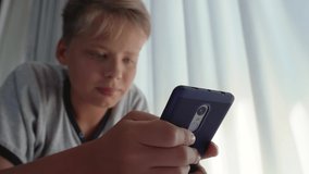 Closeup view of cute white handsome kid using modern smartphone laying in bed. Focus at phone in foreground and blurry defocused face of boy. Then focus slides to face. Real time 4k video footage.