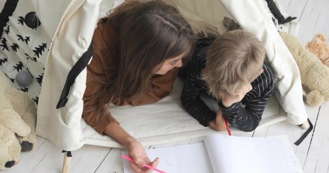 Motherhood and childhood concept. Family mom and son drawing pencils together lying in in tepee in nursery. Modern interior with wigwam, tepee, teepee. Mom spends time with child boy painting.