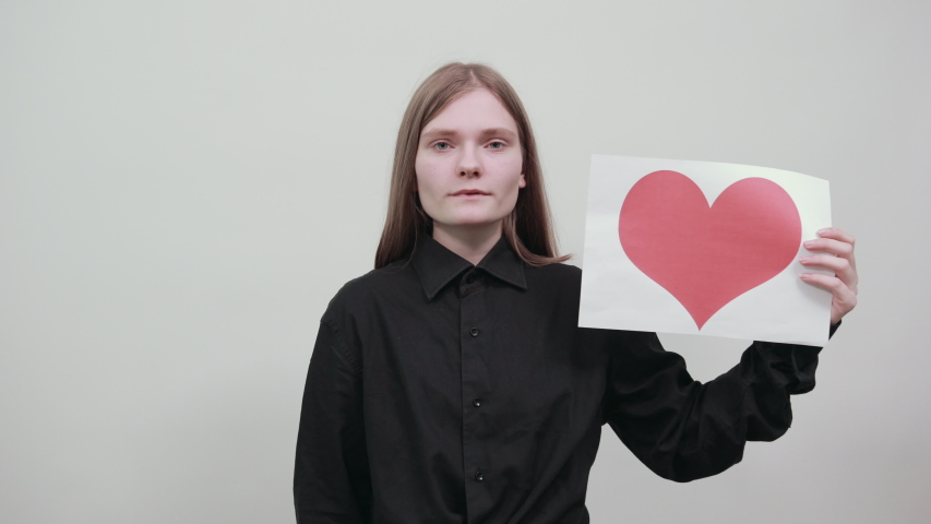 Charming caucasian young woman in fashion black shirt isolated on gray background in studio keeping paper with picture of red heart, like it. People sincere emotions, lifestyle concept.