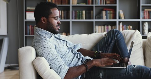 Relaxed serious millennial african ethnic guy student freelancer using laptop device leaning on sofa at home office, focused mixed race entrepreneur working distantly typing on notebook in apartment