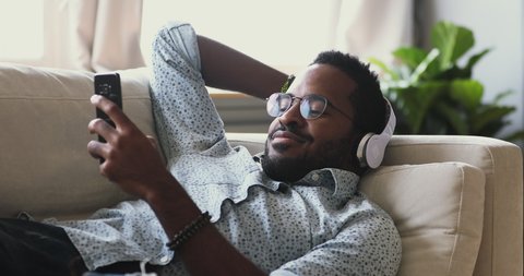 Smiling young adult mixed race african man wear wireless headphones relax on sofa listening mobile music or learning foreign language audio course in smart phone app lying resting on couch at home