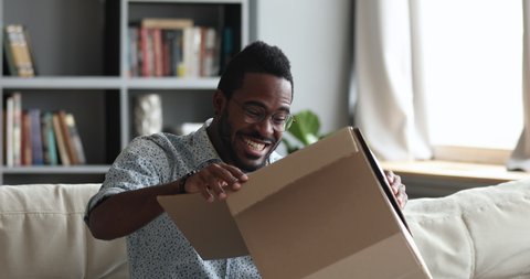Excited african male customer receive carton box with gift open post parcel sit on sofa at home, happy mixed race man consumer celebrate great retail online purchase fast shipment delivery concept