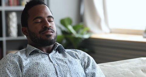 Calm relaxed young african american man meditate sit on sofa at home, serene tranquil mixed race guy doing breathing exercise take deep breath of fresh air rest with eyes closed on couch, close up