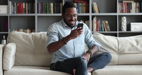 Tech addict millennial african man sitting on couch using smart phone, smiling young adult mixed race male user hipster texting sms in social media dating apps enjoy easy online communication at home