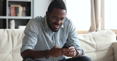 Smiling african mixed race hipster guy user holding smart phone look at cellphone using mobile social media app play game texting message enjoying easy online sport betting sitting on sofa at home