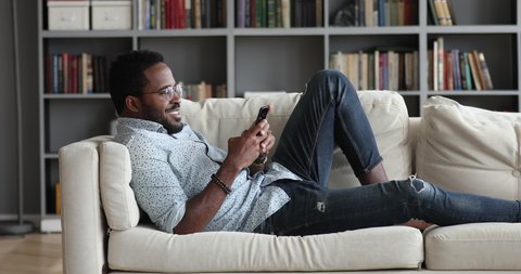 Young african american male user holding smart phone relax on sofa at home, millennial mixed race hipster guy using mobile social media apps look at cellphone enjoying leisure time with tech device