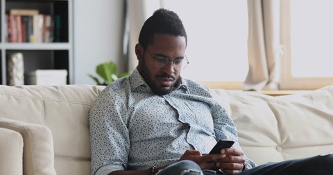 Serious young african man user holding smartphone texting message working studying in online app sit on sofa at home, focused millennial mixed race guy using cellphone surfing web on phone indoors