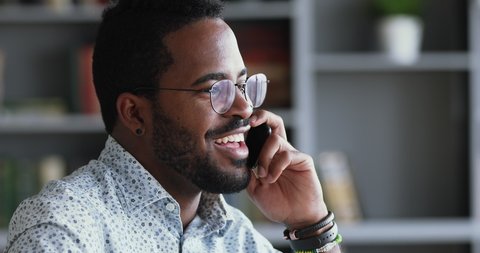 Smiling young adult mixed race professional casual businessman making business call talking on the phone in office, happy afro american salesman enjoying mobile conversation indoors, close up view
