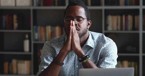 Serious african man asking for help concerned about problem or concentrating mind at work, religious mixed race male student worker sit with eyes closed put hands in prayer praying with hope concept