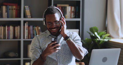 Smiling young african business man student professional wearing headphones listen to mobile music playing in smartphone app, happy mixed race guy relaxing at work desk using phone at home office